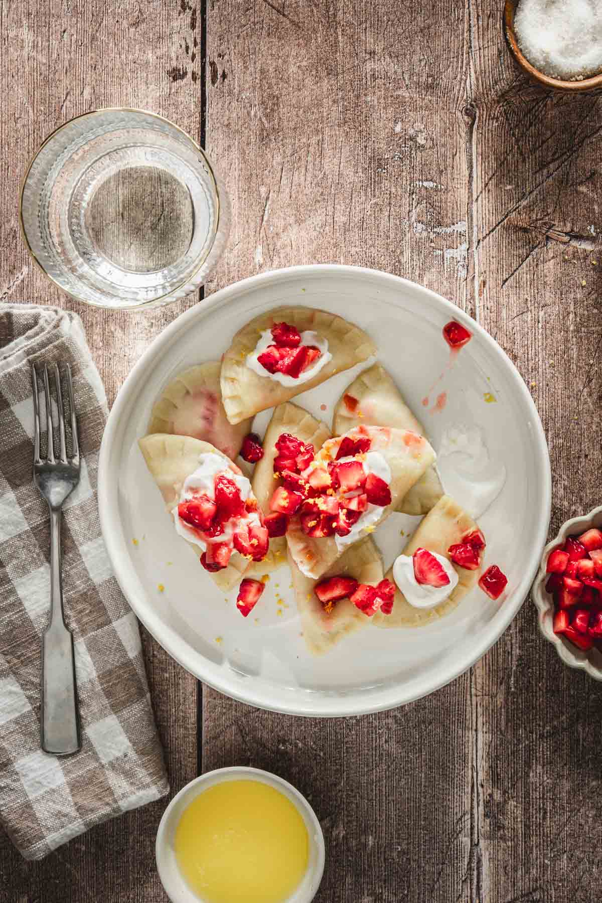 cooked pierogis in a serving bowl, topped with fresh cut up strawberries, napkin, fork, glass with water, dish with fresh cut up strawberries