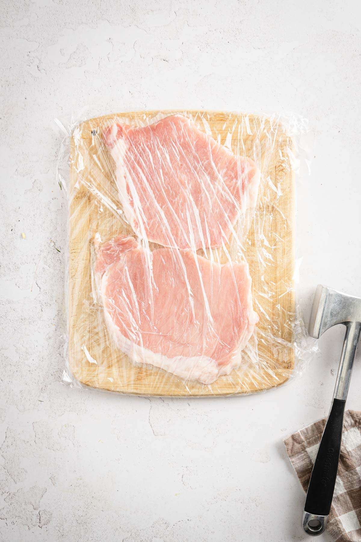 two pork chops on a cutting board, covered with plastic wrap