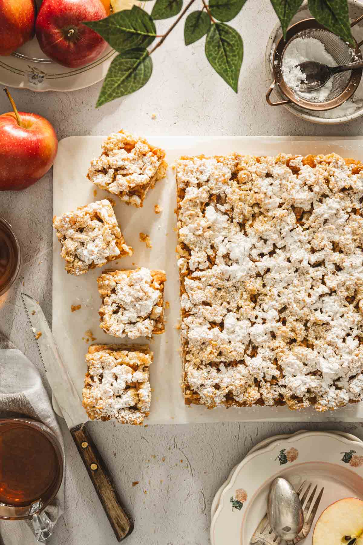 szarlotka on a serving stone tray with powdered sugar applied, knife, few serving plates, bowl with powdered sugar, glasses with tea, plate with apples, four pieces cut out