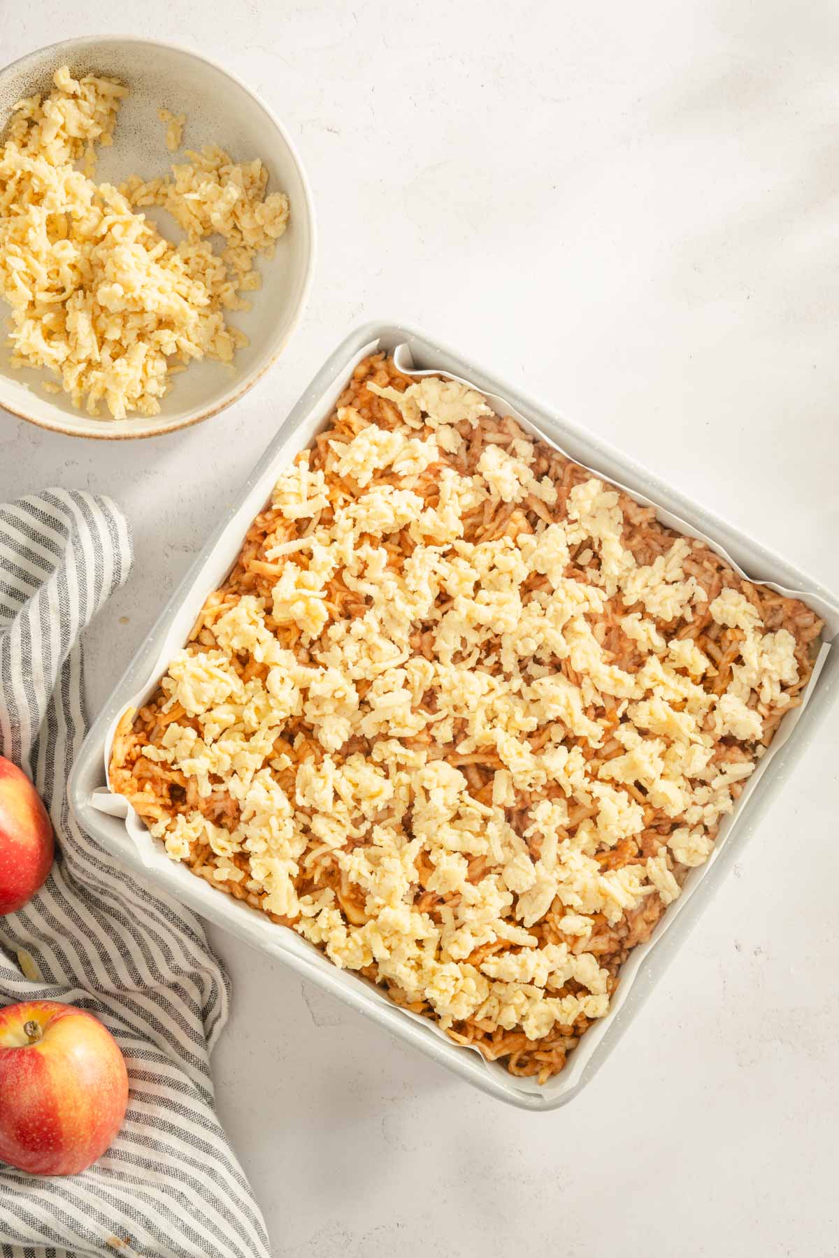 baking dish with Szarlotka ready to bake, linen, whole apple, small bowl with little shredded dough