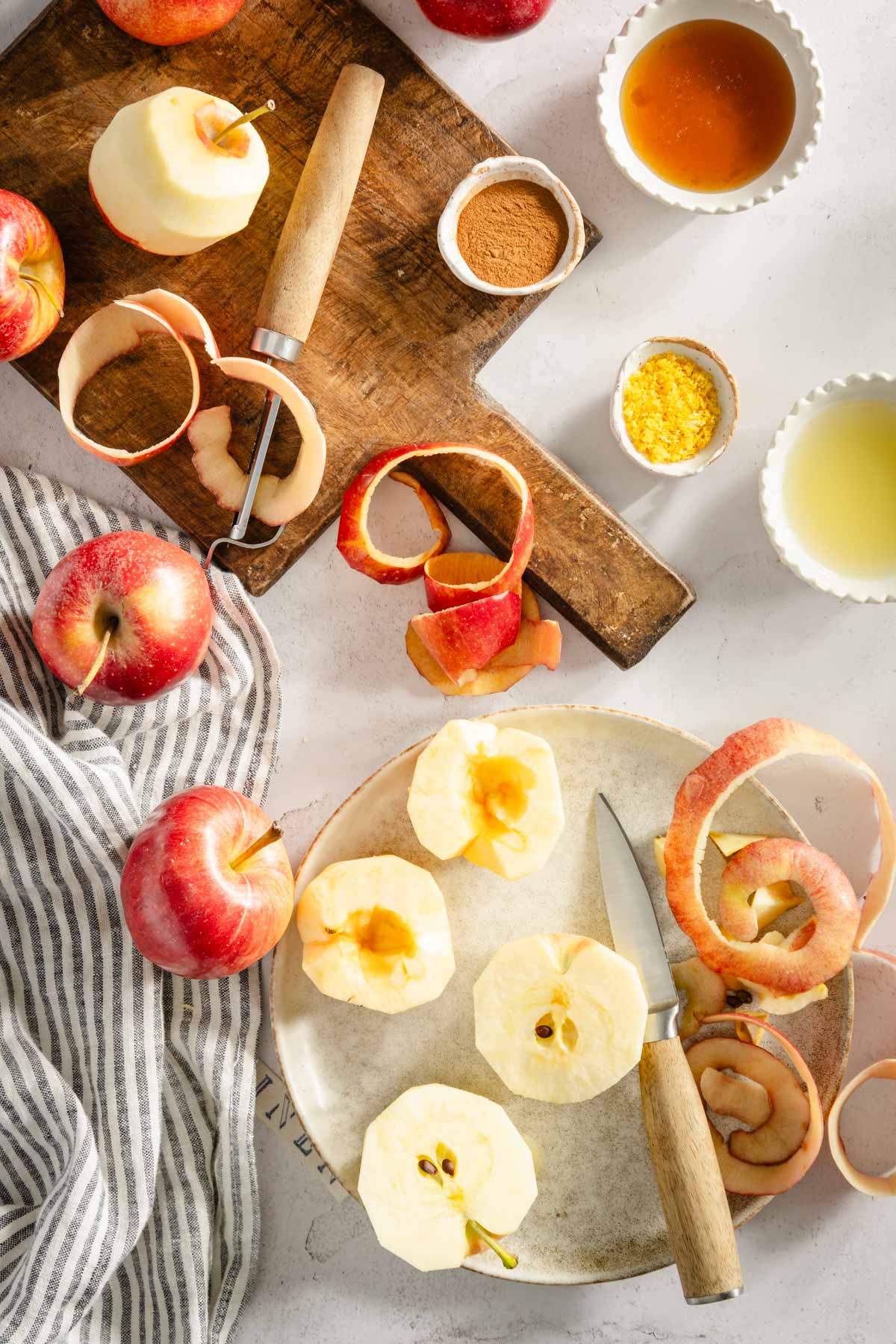 apples on a cutting board, some peeled, small knife and a peeler, small bowls with other ingredients