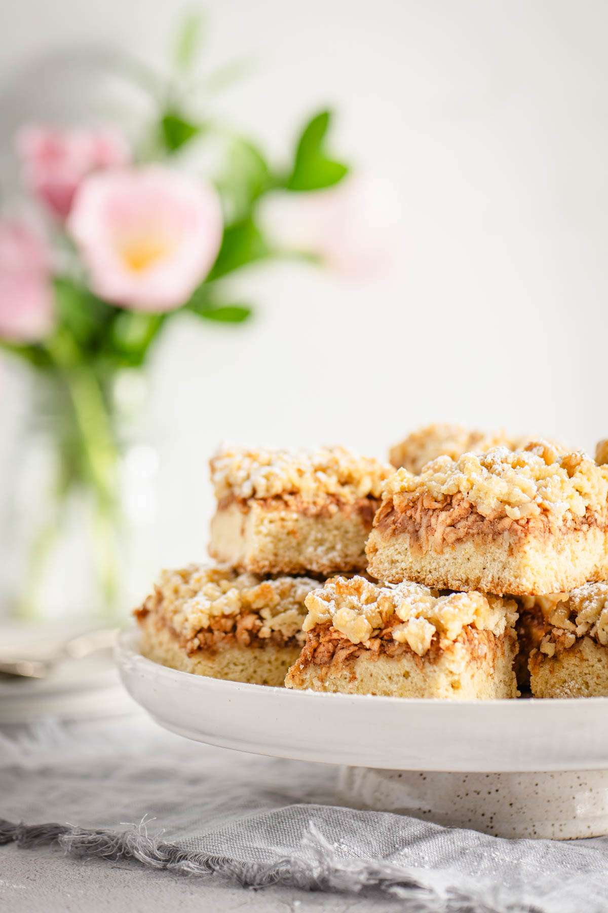 Slices of Szarlotka on a serving plate, vase with flowers in the background