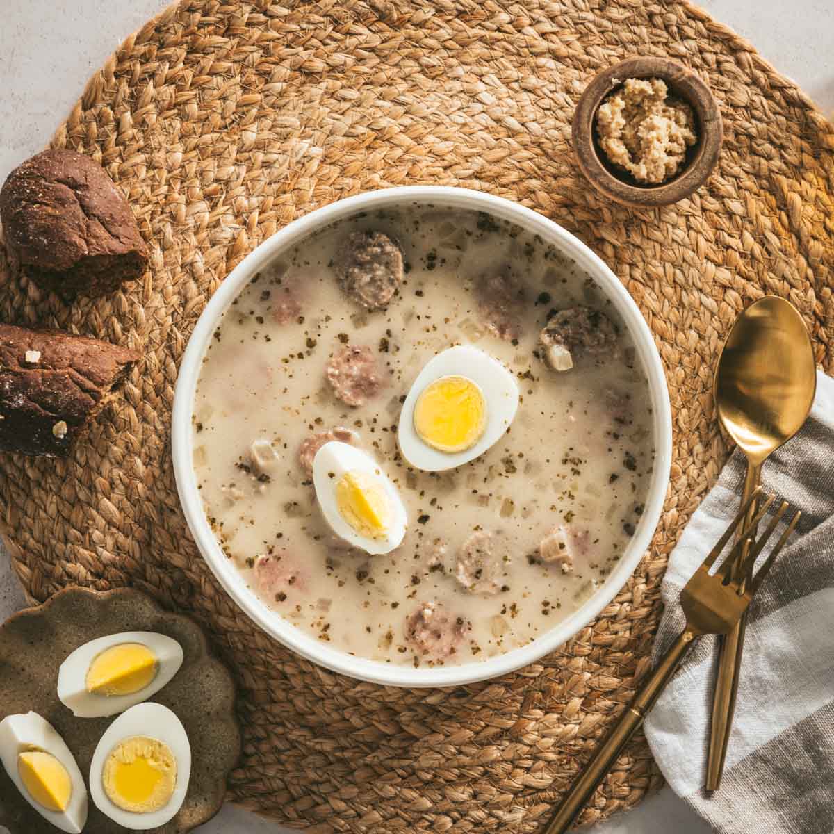 Zurek Soup in a serving bowl with hard boiled egg, piece of bread, small dish with horseradish, spoon, fork, cut up egg on a small plate