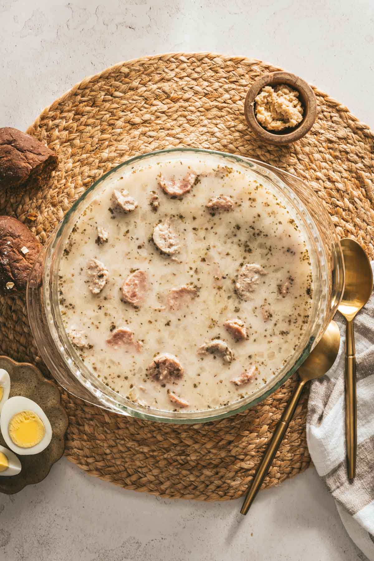 Zurek Soup in a glass soup bowl, piece of bread, small dish with horseradish, spoon, fork, cut up egg on a small plate, bowl with cut up eggs