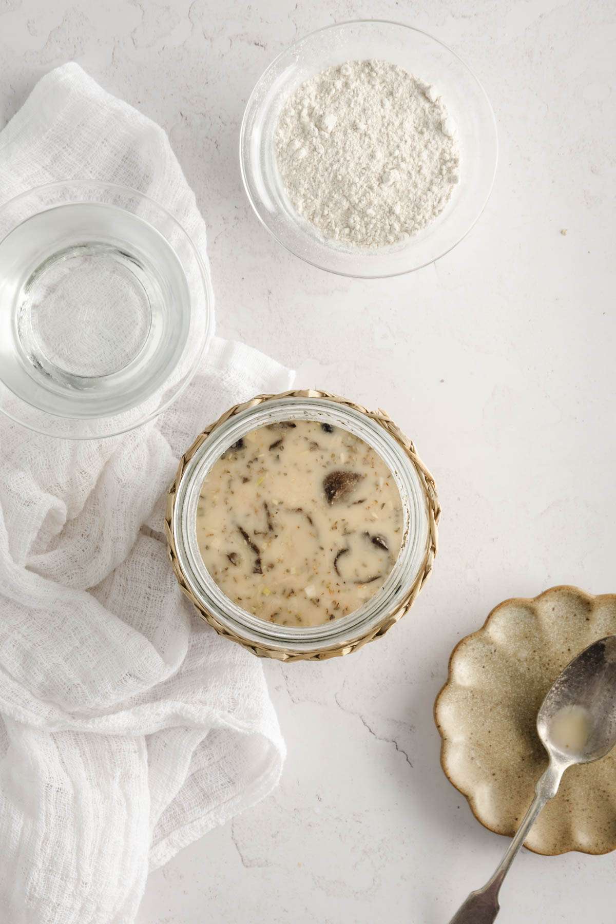 top view of jar with zakwas, spoon for mixing, two small bowls , one with water, one with flour, fermented rye starter