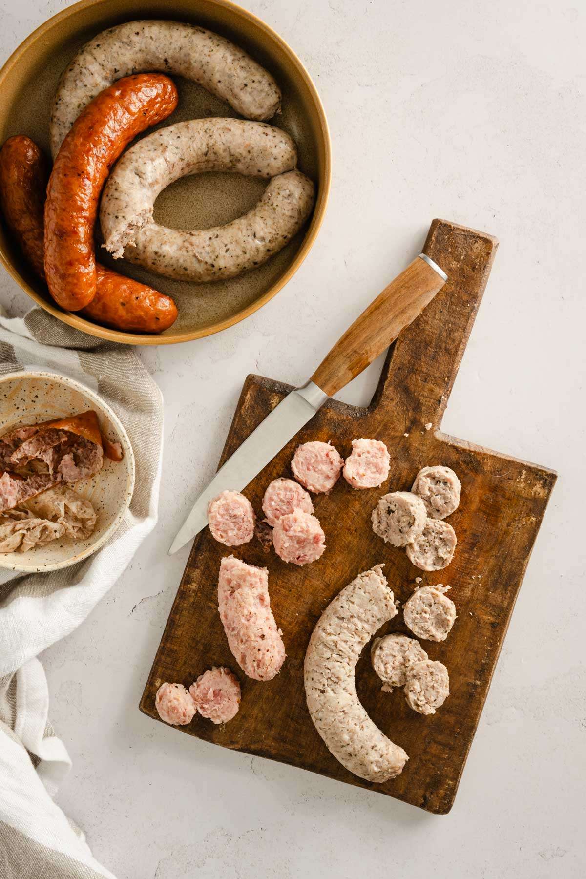 cutting board with sausages, dish to the side with whole sausages, small dish with peeled off sausage skin, small knife