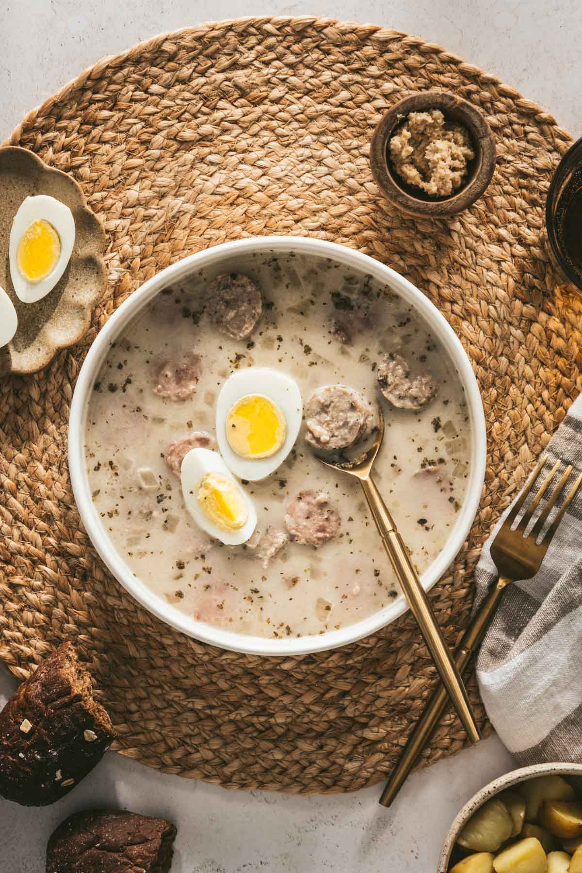 Zurek Soup in a serving bowl with hard boiled egg, piece of bread, small dish with horseradish, spoon, fork, cut up egg on a small plate