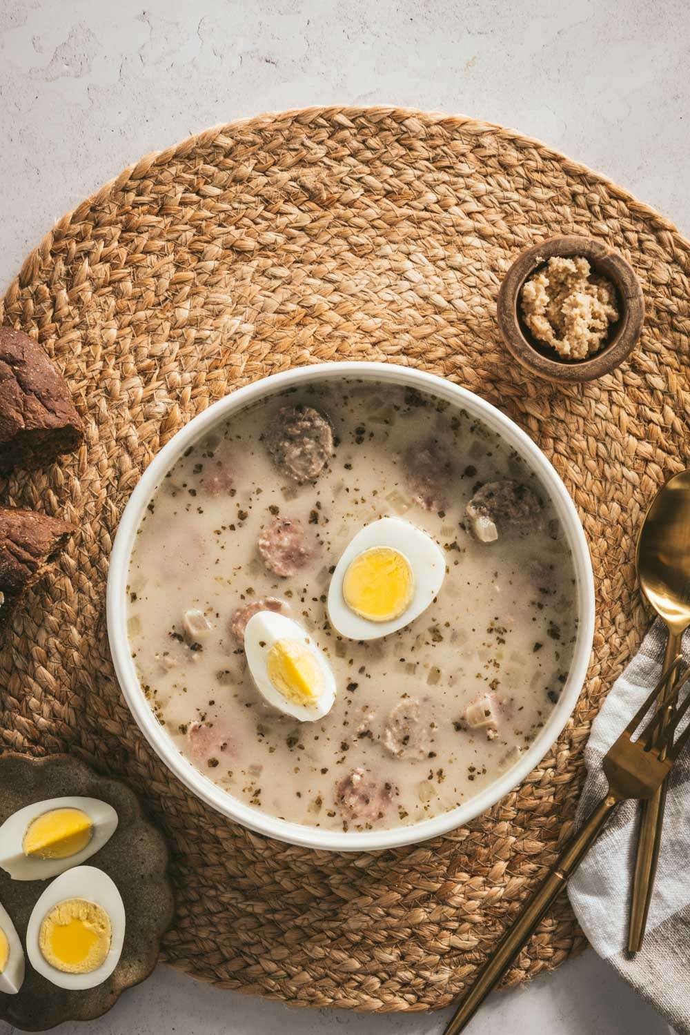 Zurek Soup in a serving bowl with hard boiled egg, piece of bread, small dish with horseradish, spoon, fork, cut up egg on a small plate