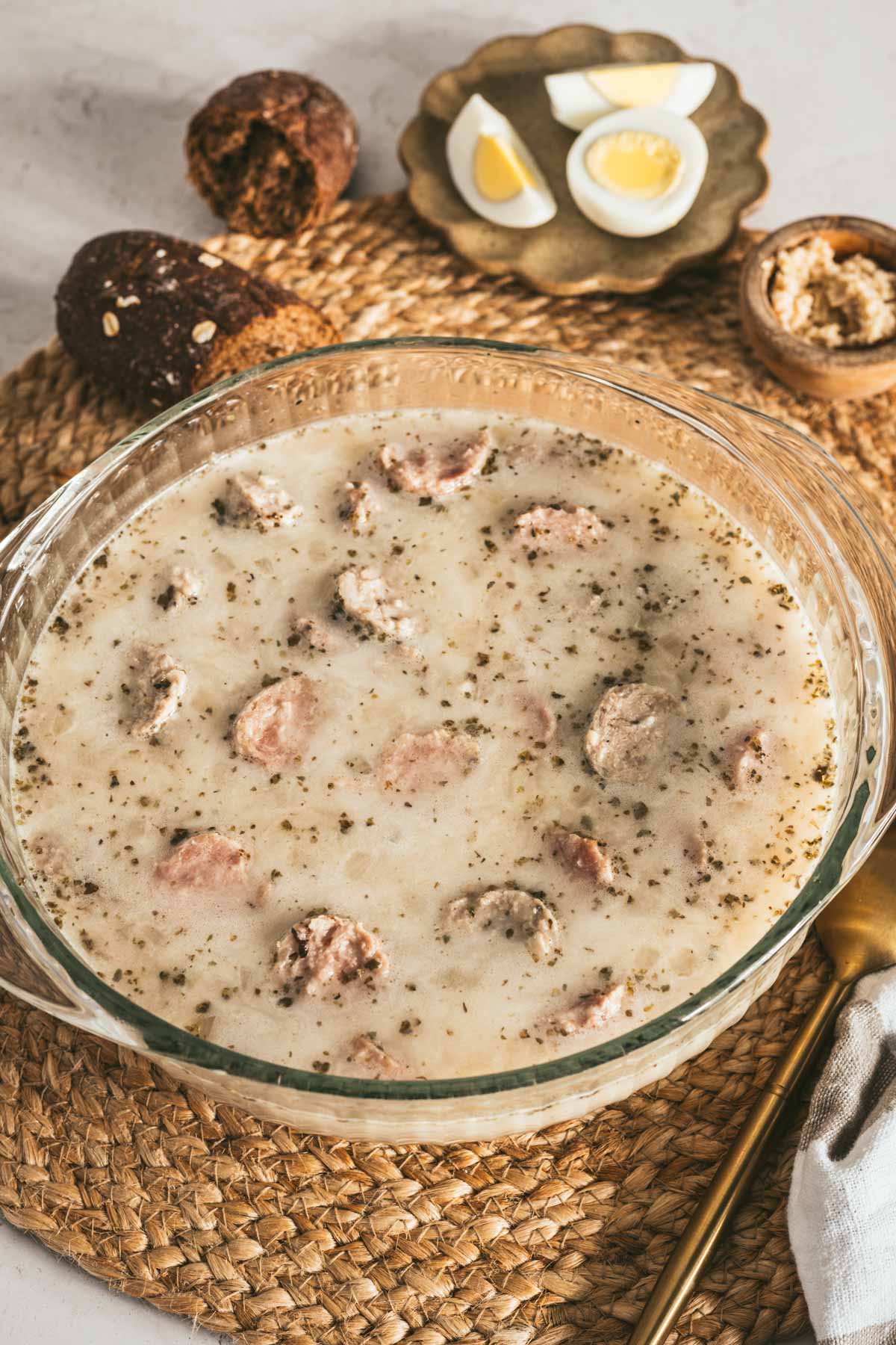 Zurek Soup in a glass soup bowl, piece of bread, small dish with horseradish, spoon, fork, small plate with cut up egg