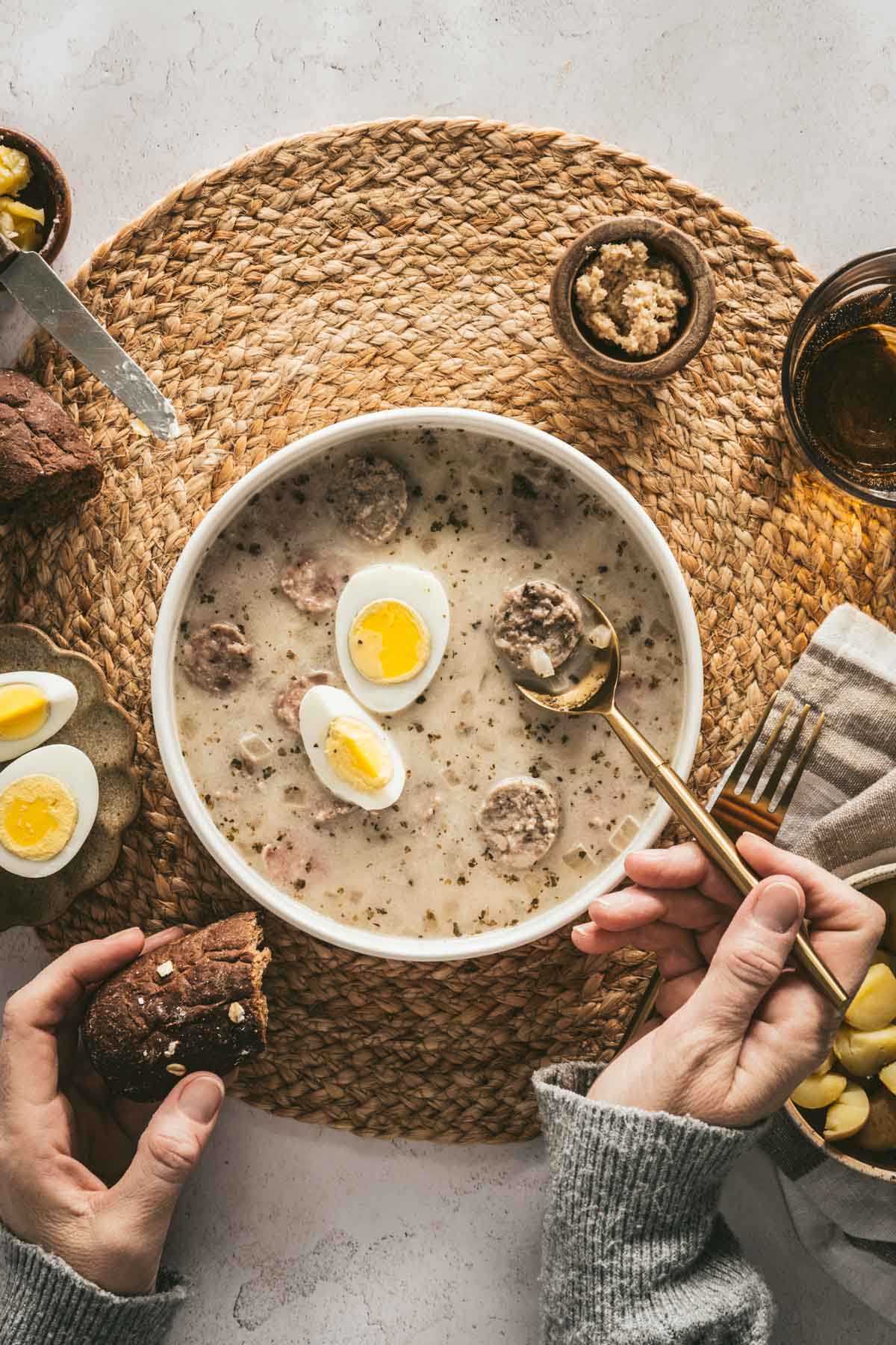 Zurek Soup in a serving bowl with hard boiled egg, piece of bread, small dish with horseradish, spoon, fork, cut up egg on a small plate, hands in frame holding spoon and piece of bread