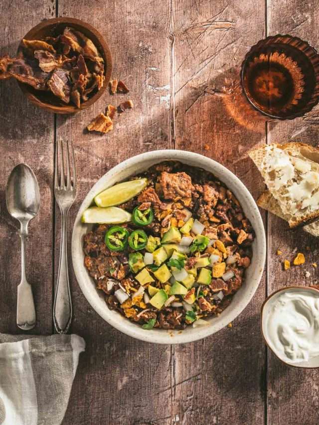 chili in a serving bowl garnished with diced avocado, limes, jalapenos, utensils on the left, glass of water, bowl with bacon, sour cream, two slices of bread, napkin