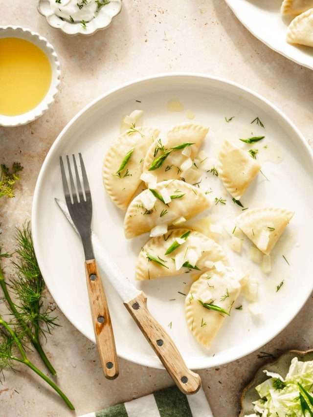 plate with pierogis garnished with fried onion and chives, side salad, dish with cooked onions, platter with cooked pierogis, dish with melted butter and yogurt