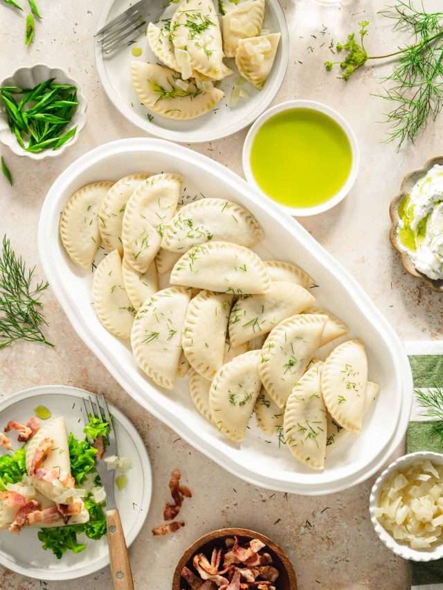 dill pierogis arranged on a oval platter, two plates with pierogis, dish with green onions, fried bacon, cooked onions and dill oil