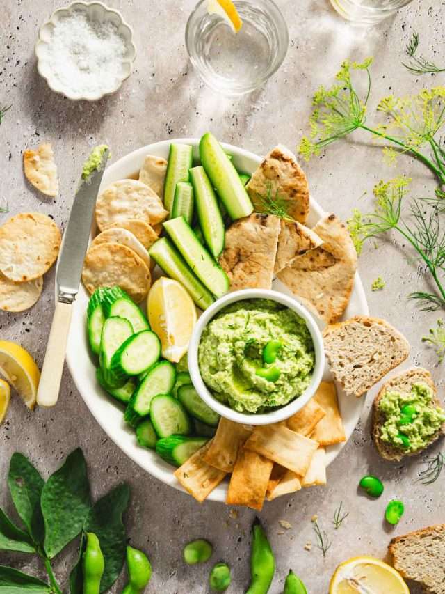 platter with veggies, bread and crackers and a bowl with fava bean hummus; lemon slices, glass of water, bread