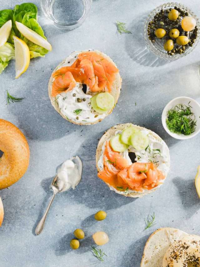 pieces of salmon on a bagel with cream cheese and sliced cucumbers, dish with olives, fresh dill; glasses with water