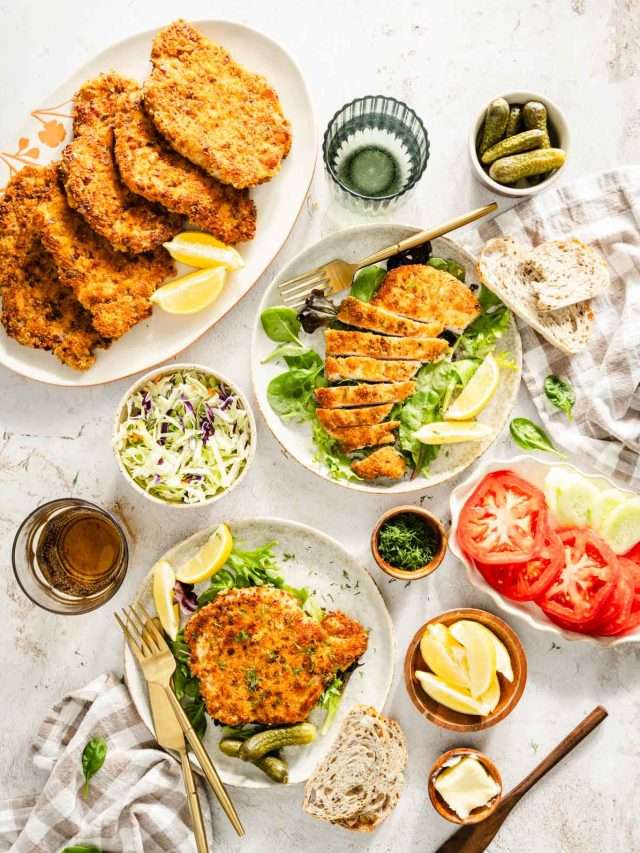 cooked por chops presented on plates with salad, dish with fresh dill, glass of water, pickles, utensils and slices of bread, dish with salad and sliced tomatoes