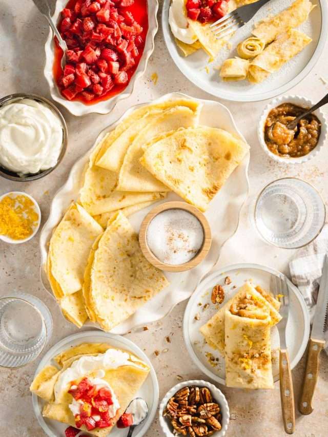nalesniki styled on a serving plate, small dishes with sour cream, apple sauce, sugar. Dish with cut up strawberries. Various plates with nalesniki folded in different ways