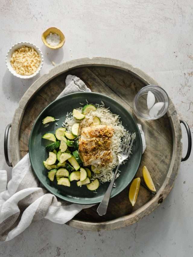 pistachio crusted cod on a bed of sauté zucchini and white rice; lemon wedges and small dish with cheese and salt on the side; all on wooden serving tray