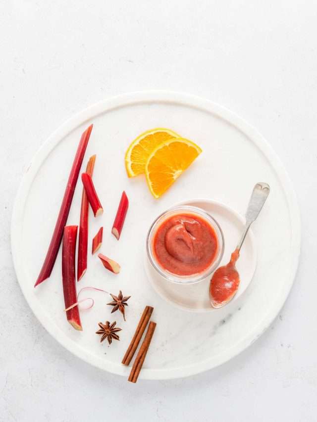 rhubarb butter in a small jar, placed on white marble tray, few rhubarb stalks, cinnamon sticks and orange slices, spoon