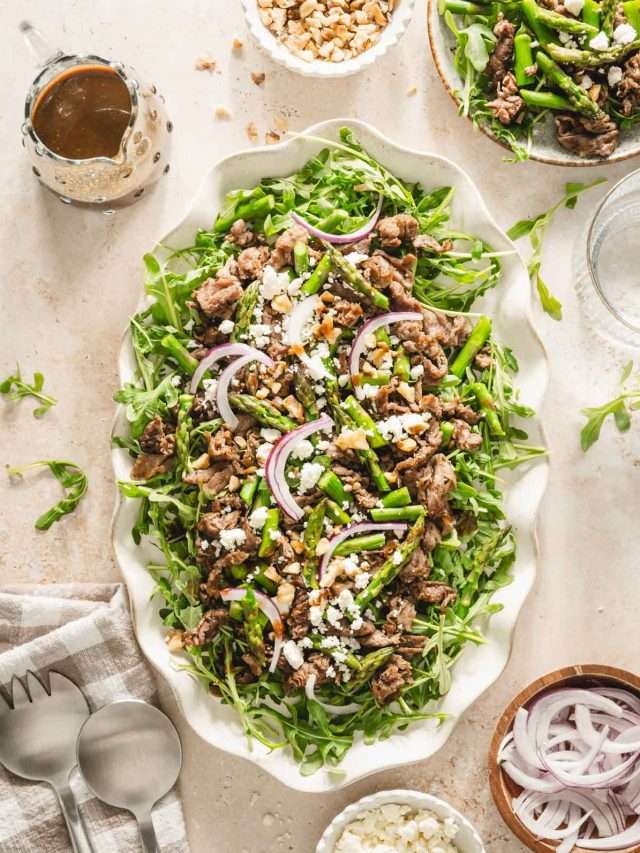 shaved beef and asparagus salad on a serving platter, utensils, small dishes with feta cheese, onions, walnuts, glass of water and glass with vinaigrette