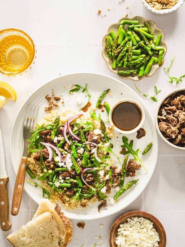 shaved beef and asparagus salad on a serving plate, utensils, small dish with onions, walnuts, meat, asparagus and dressing, slices of bread, glass with water, lemon wedges