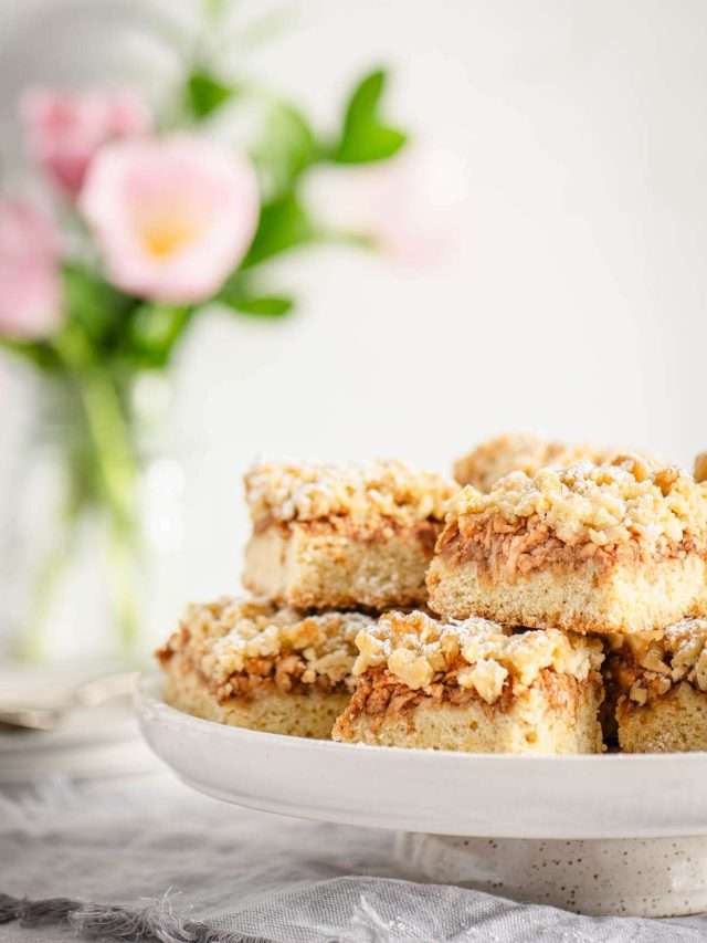 Slices of Szarlotka on a serving plate, vase with flowers in the background